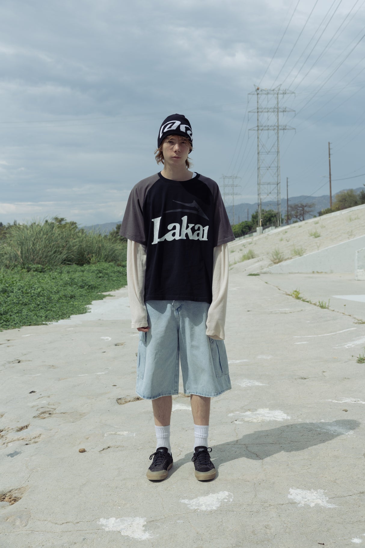 Person wearing a black 'Laka' t-shirt and light blue shorts standing on a road with power lines in the background.