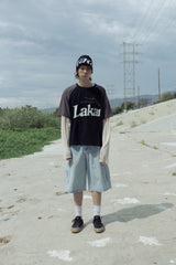 Person wearing a black 'Laka' t-shirt and light blue shorts standing on a road with power lines in the background.